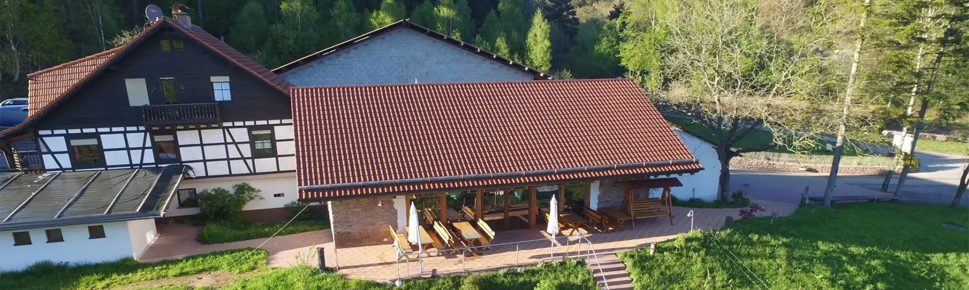 Fachwerkhaus mit rotem Ziegeldach und überdachter Terrasse mit Tischen und Stühlen, umgeben von Wiesen und Bäumen im Odenwald, Hilsighof.