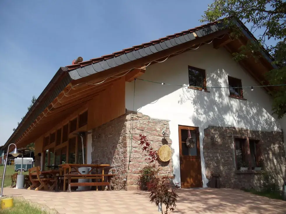 Haus mit weißer Fassade, Natursteinmauerwerk und braunem Satteldach, davor Terrasse mit Holztisch und Bänken.