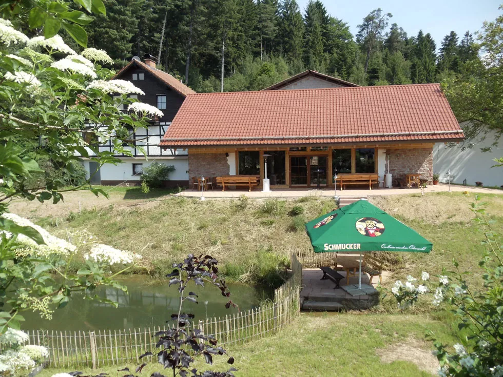 Haus mit rotem Ziegeldach vor Wald, davor Teich mit grünem Sonnenschirm und Sitzbereich auf Holzsteg