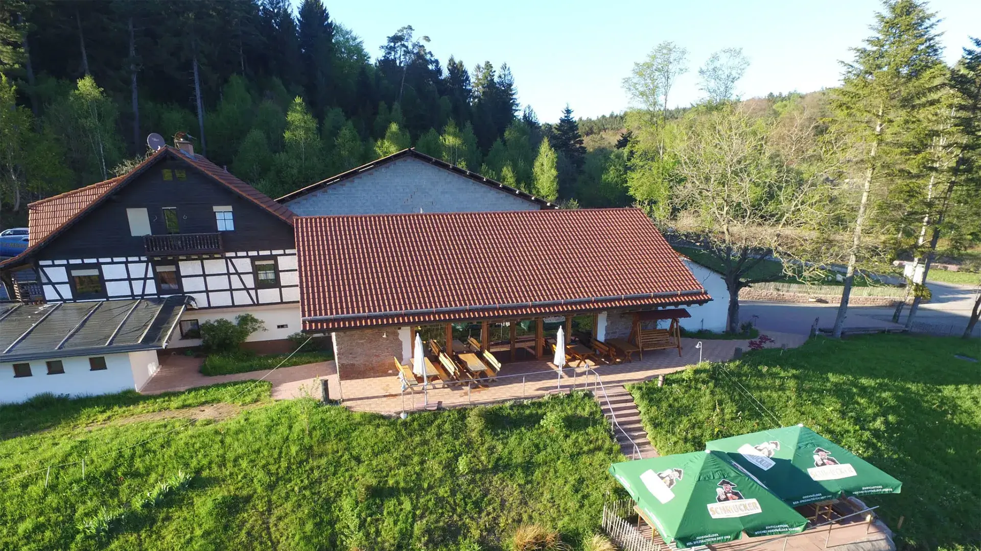 Fachwerkhaus mit rotem Ziegeldach und überdachter Terrasse mit Tischen und Stühlen, umgeben von Wiesen und Bäumen im Odenwald, Hilsighof.