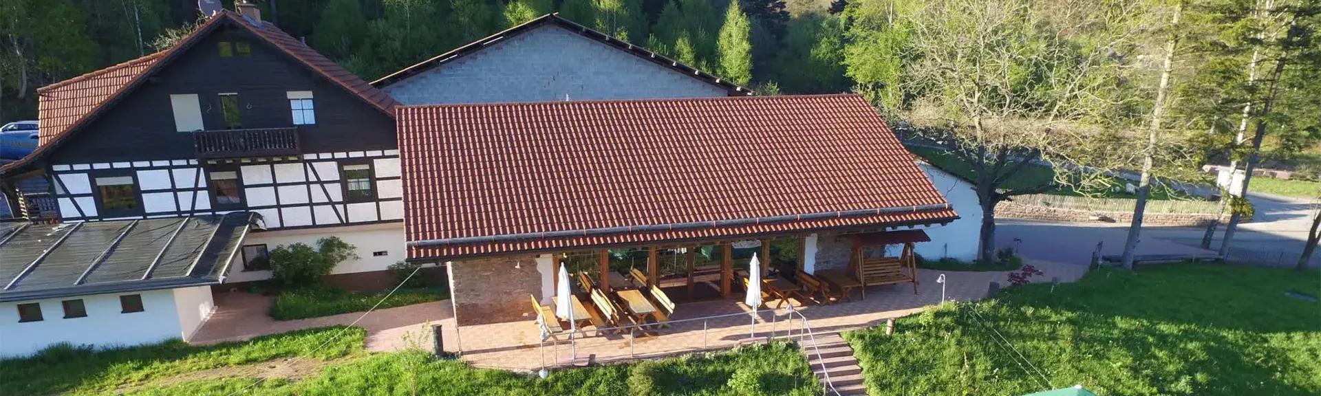 Hilsighof Vogelperspektive Fachwerkhaus mit rotem Ziegeldach und überdachter Terrasse mit Tischen und Stühlen, umgeben von Wiesen und Bäumen im Odenwald, Hilsighof.