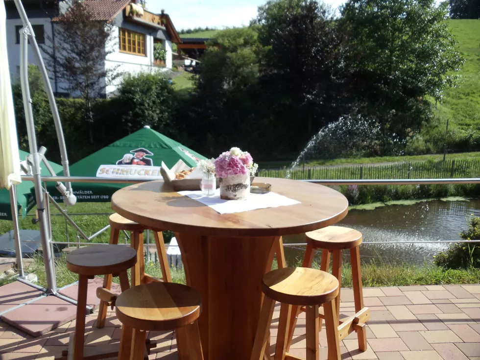 Runder Holztisch mit fünf Hockern auf Terrasse neben Teich, Tisch mit Blumenarrangement und Servietten.
