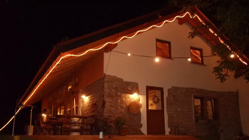Haus mit Satteldach und Steinmauer, beleuchtet von Außenlampen und einer Lichterkette am Dachrand bei Nacht.